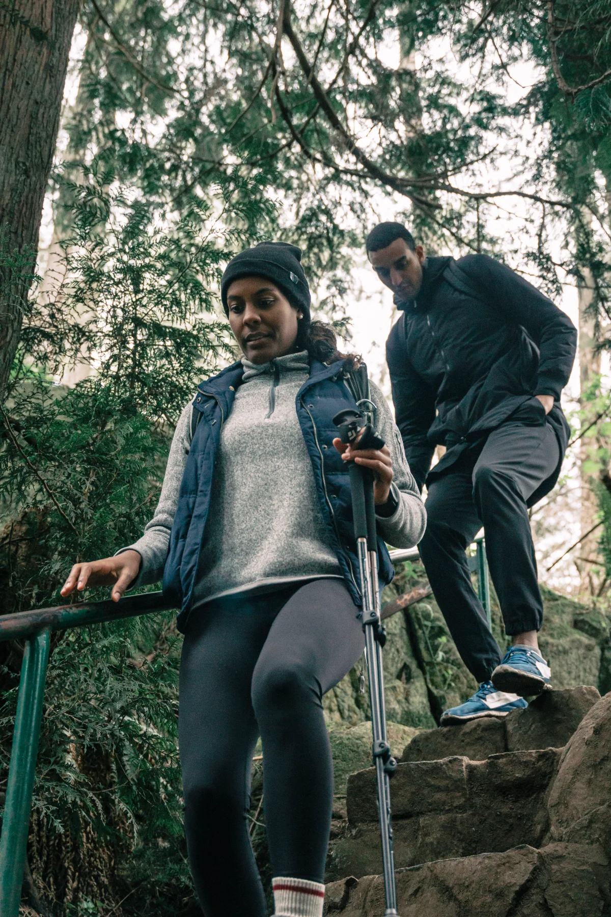 A woman hiking down a forest trail with trekking poles, a friend just behind her