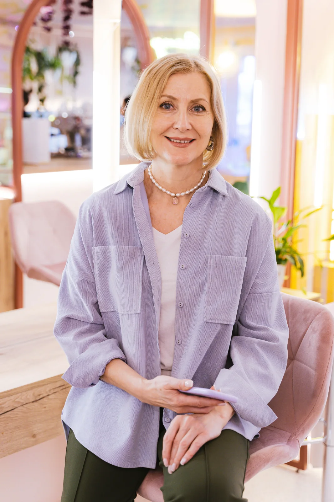 A smiling woman with a blonde bob and pearl necklace, seated with her phone