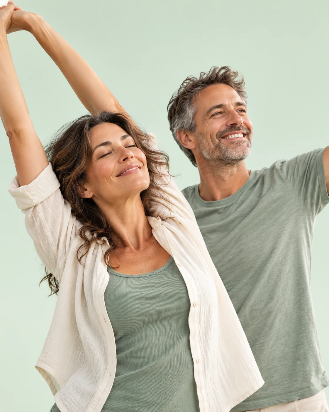 A smiling woman stretching her arms overhead beside a smiling man, against a soft mint background